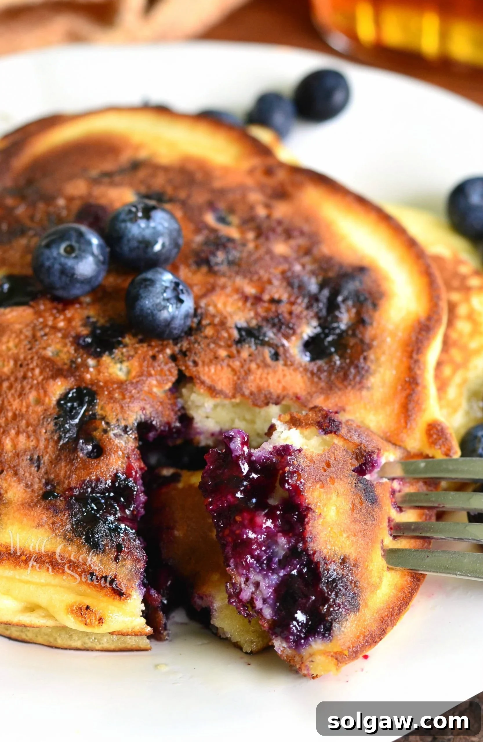 Single serving buttermilk pancakes topped with syrup and blueberries on a white plate with a taking one bite out of the pancakes.