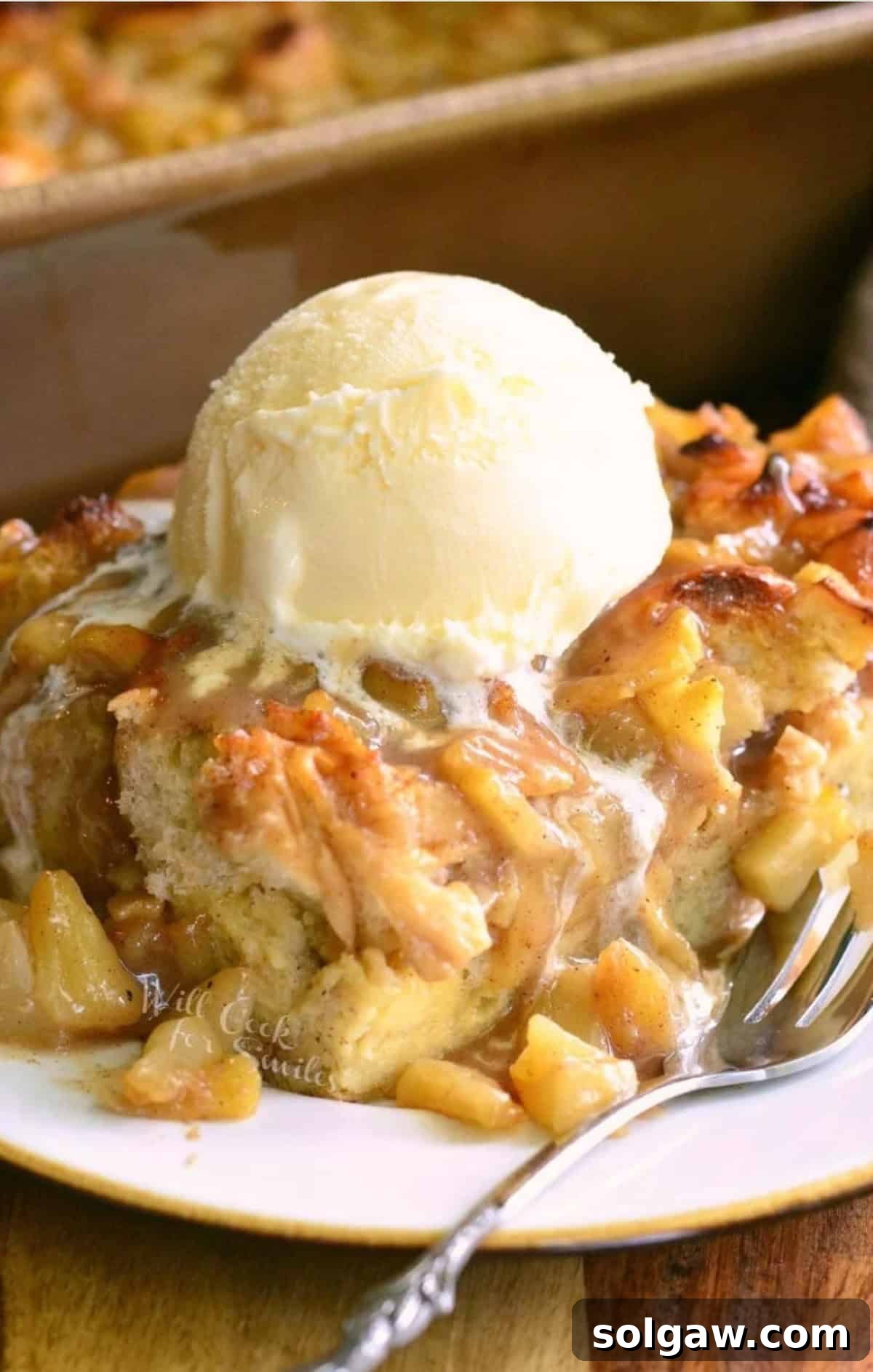 A slice of warm apple pie bread pudding topped with melting vanilla ice cream on a white plate, ready to be enjoyed.