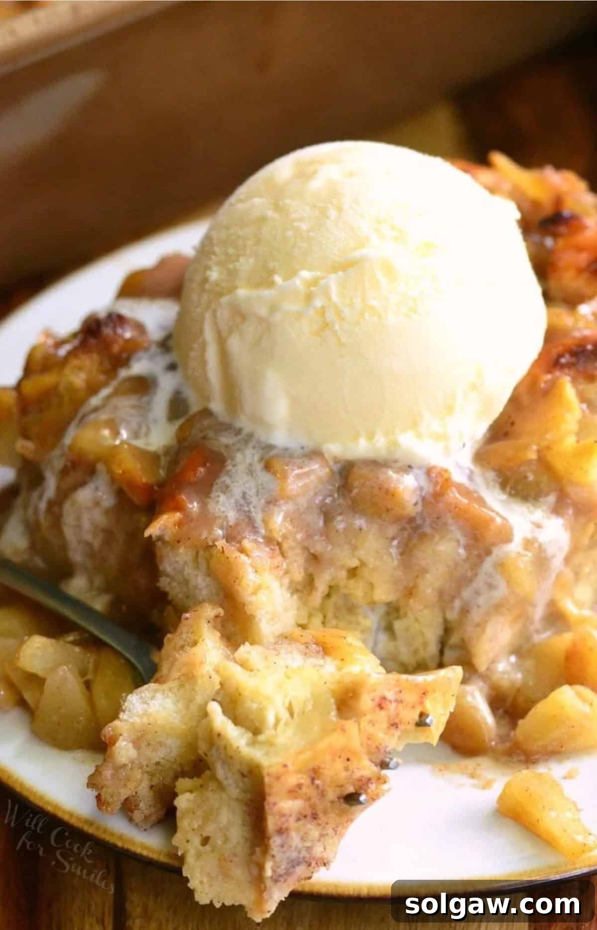 A close-up of a slice of apple pie bread pudding with a scoop of vanilla ice cream, showing a fork having taken a bite out.