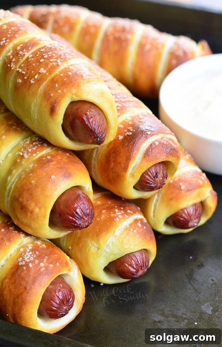Pretzel Wrapped Hot Dogs stacked up on baking sheet with creamy Maple Dijon Dipping Sauce in a white bowl