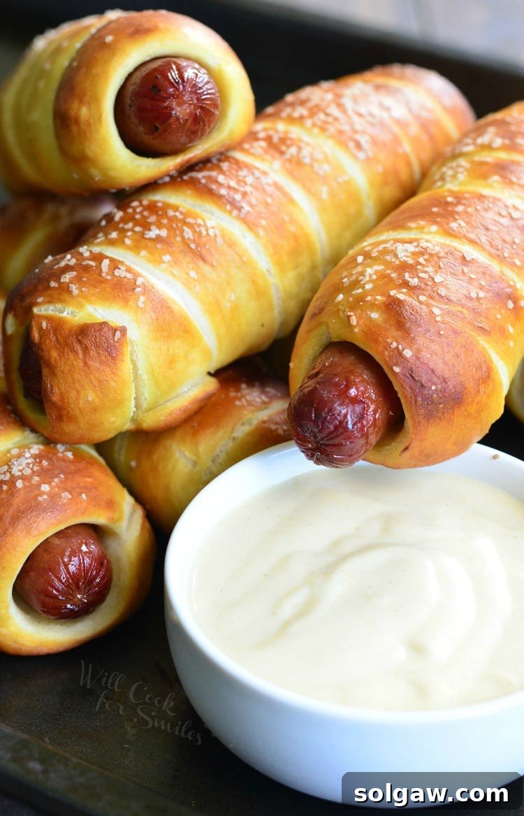 Pretzel Wrapped Hot Dogs stacked on a baking sheet with creamy Maple Dijon Dipping Sauce in a white bowl, ready to be served