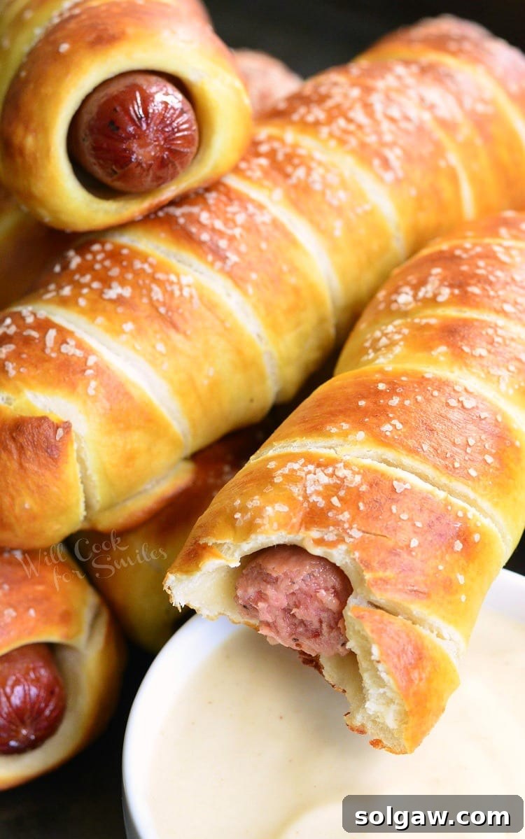 Pretzel Wrapped Hot Dogs stacked on a baking sheet, one with a bite taken out, showcasing the hot dog inside, alongside a bowl of Maple Dijon Dipping Sauce