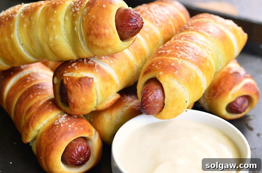 Batch of freshly baked Pretzel Wrapped Hot Dogs arranged on a baking sheet with a small white bowl of Maple Dijon Dipping Sauce