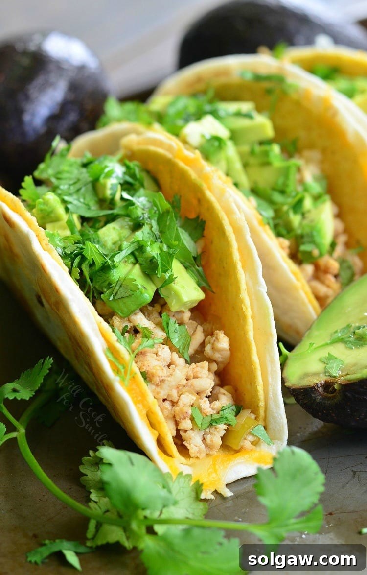 3 avocado turkey tacos on a slate table with avocados in the background and to the left of the tacos as viewed close up.