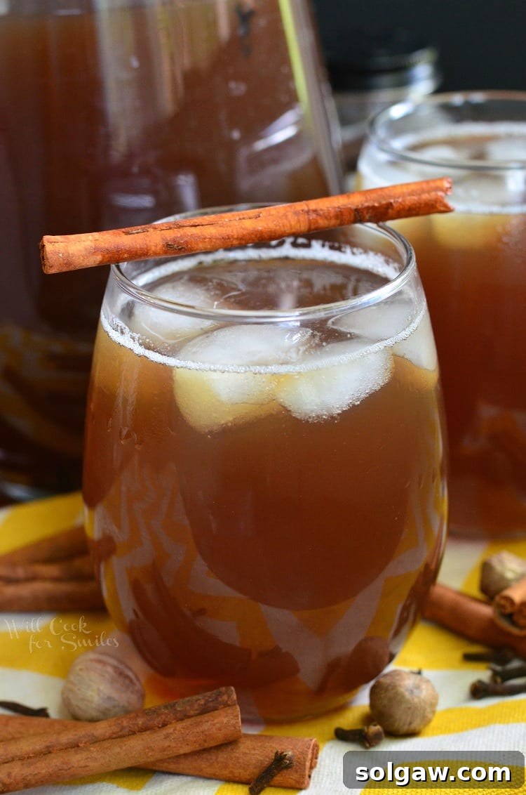 A glass of spiced iced tea with ice and a cinnamon stick, surrounded by whole nutmeg, cloves, and cinnamon sticks on a yellow and white tablecloth.