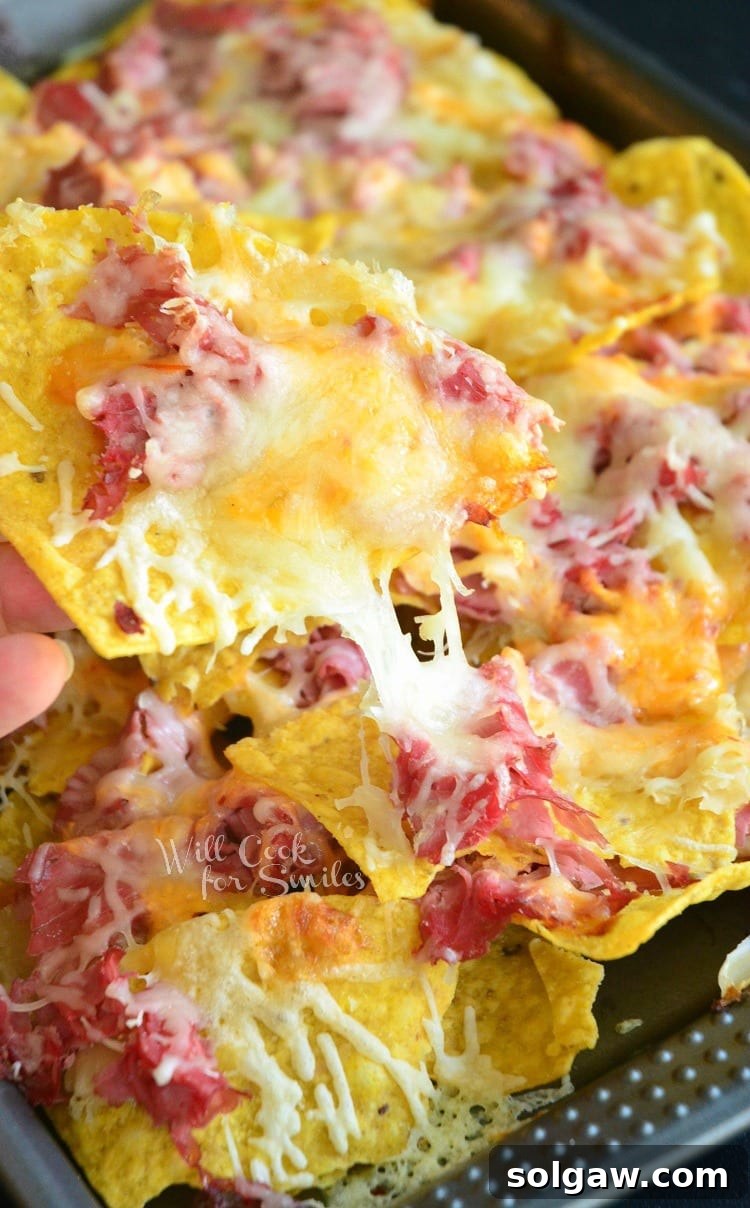 Lifting a tortilla chip loaded with corned beef, sauerkraut, and melted cheese off a baking sheet – a perfect bite of Reuben Nachos