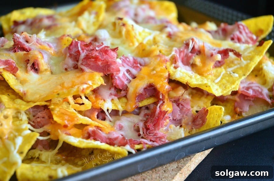 Horizontal shot of golden tortilla chips topped with corned beef and melted Swiss cheese on a baking sheet, ready for more toppings