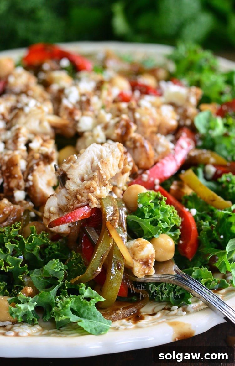 A vibrant kale salad topped with perfectly sautéed balsamic chicken and colorful vegetables, chickpeas, and generous amounts of crumbled feta cheese, served on a pristine white plate with a fork ready to enjoy.