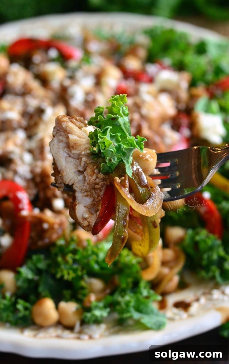 A close-up shot of a fork lifting a bite of balsamic chicken, sautéed vegetables, and chickpeas from a fresh kale salad, all adorned with crumbled feta cheese on a white plate.
