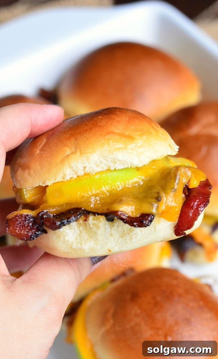 A hand holding an Apple Bacon Cheddar Baked Slider, with more sliders visible in the background in a baking dish.
