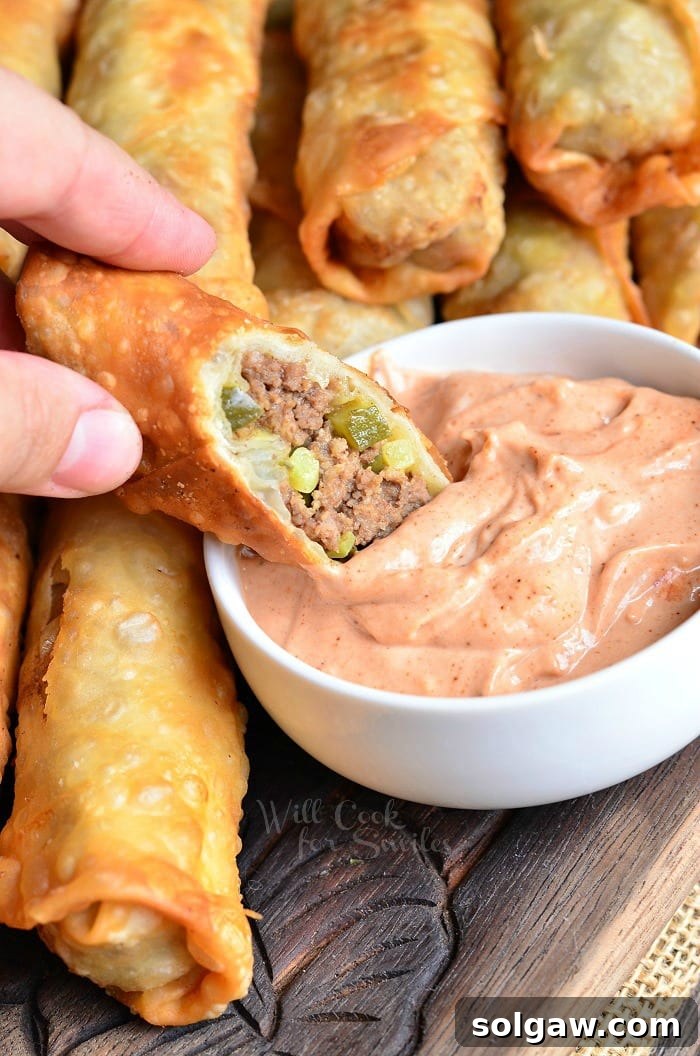 Cheeseburger Egg Rolls being dipped into a white bowl of sauce, ready to be eaten.