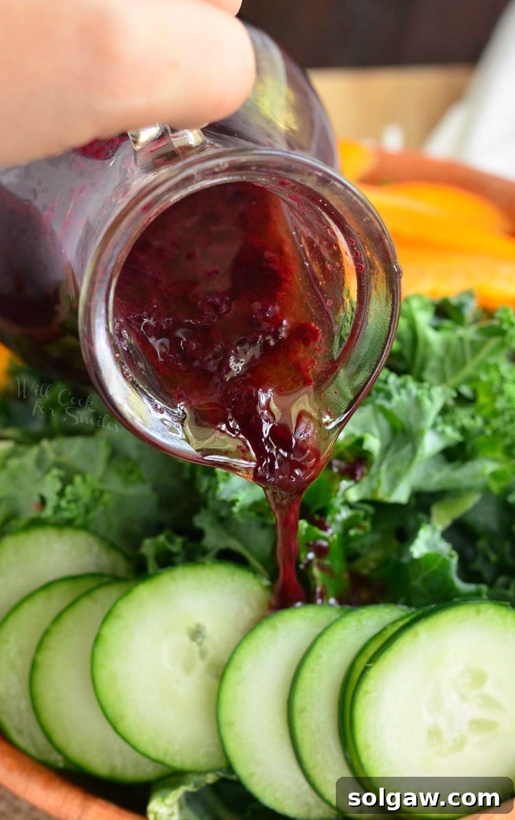 A close-up of fresh salad greens and vegetables being generously drizzled with homemade Blueberry Lavender Vinaigrette from a glass container, showcasing the dressing's vibrant color and consistency.