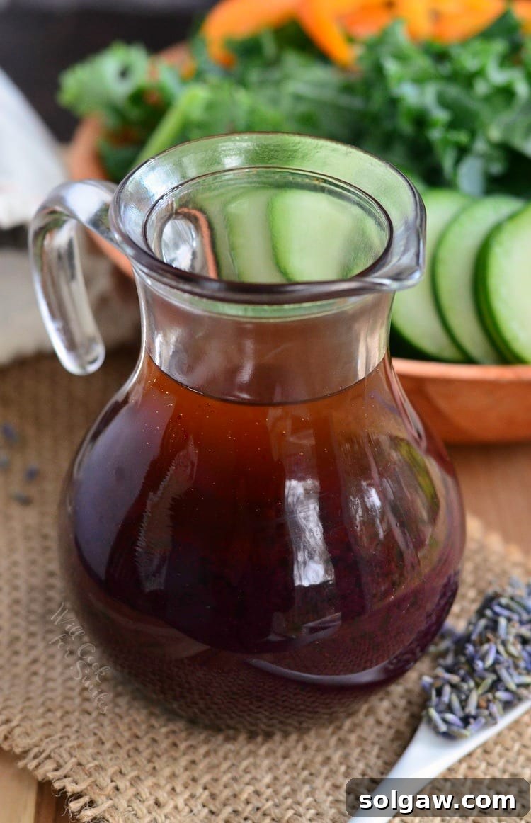 A beautifully composed shot of Blueberry Lavender Vinaigrette in a glass jar, accompanied by a spoonful of lavender, with a salad waiting in the background.