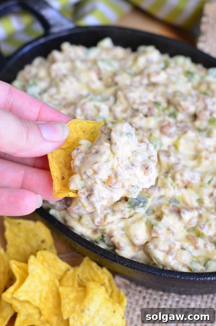 A tortilla chip being dipped into the warm, gooey Cheesy Sausage Peppers and Onions Dip, showing its creamy texture