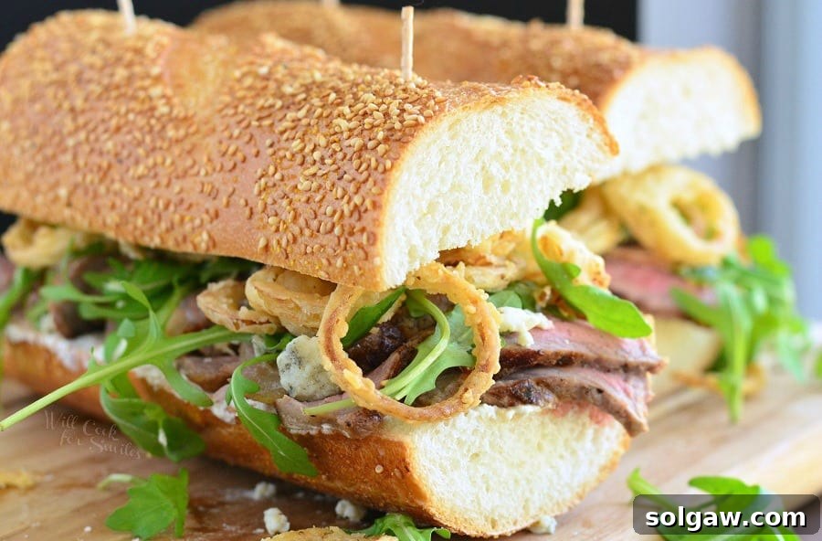 Close-up of a stacked Steak Sandwich, highlighting the crispy homemade onion rings, blue cheese crumbles, and fresh arugula