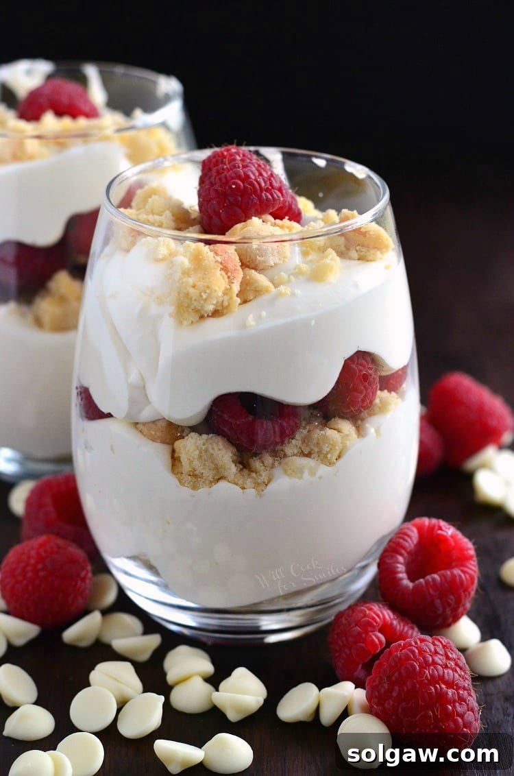 Elegant Raspberry White Chocolate Mousse Parfait in a stemless wine glass, surrounded by white chocolate chips and fresh raspberries on a pristine white table