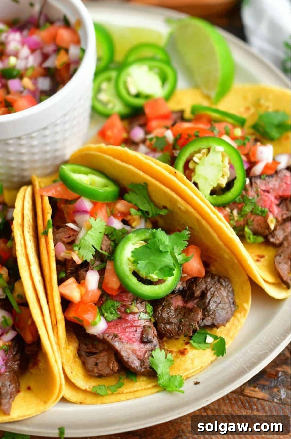 Close-up of three mouthwatering Carne Asada Tacos, generously topped with fresh Pico de Gallo and vibrant green cilantro, served on a light brown plate with additional lime wedges and a bowl of salsa.