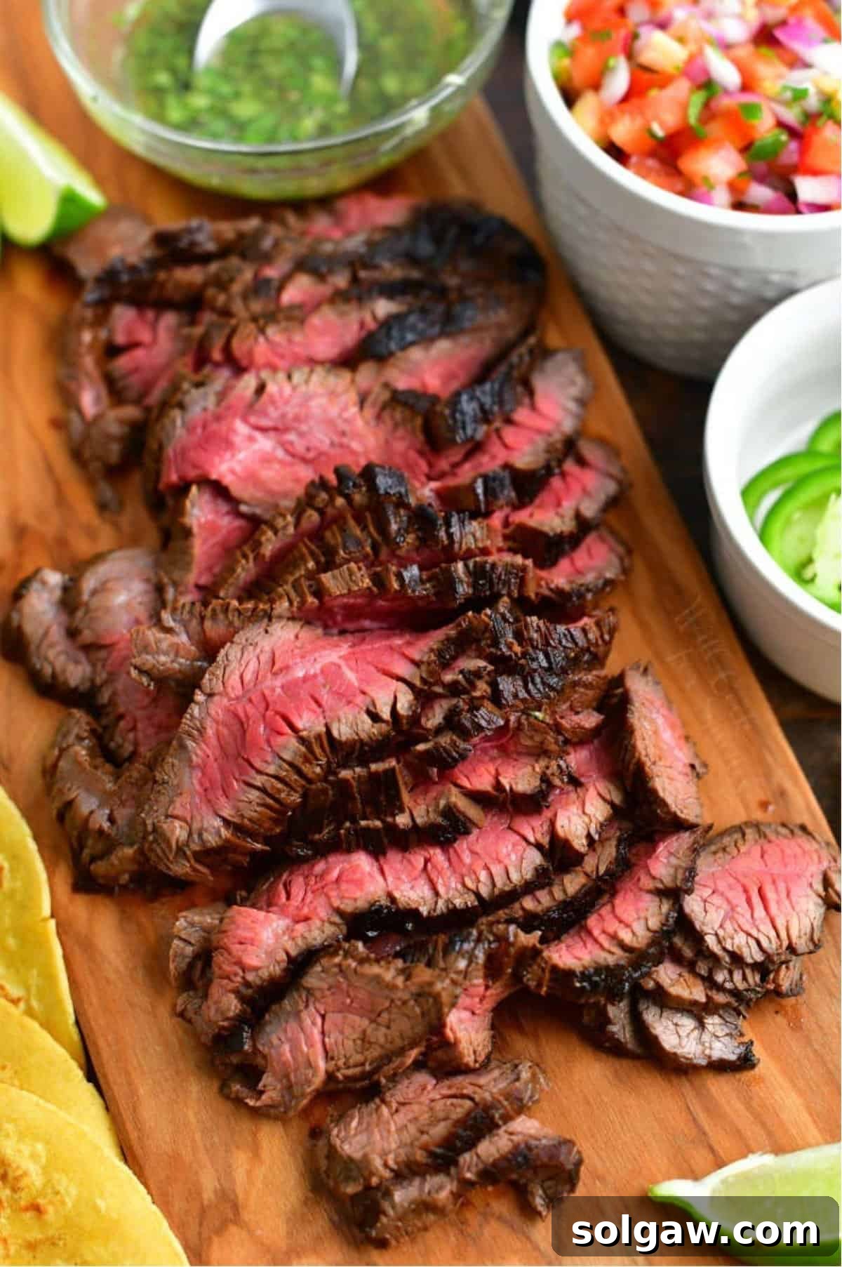 Perfectly sliced carne asada steak arranged on a wooden cutting board, alongside vibrant Pico de Gallo and fresh cilantro for assembling delicious tacos.