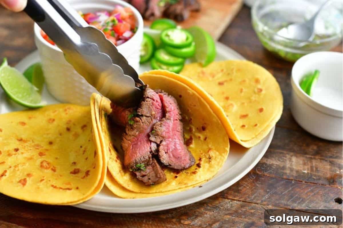 A hand is shown adding perfectly grilled and sliced carne asada steak into warm taco shells, preparing them for a delicious meal.