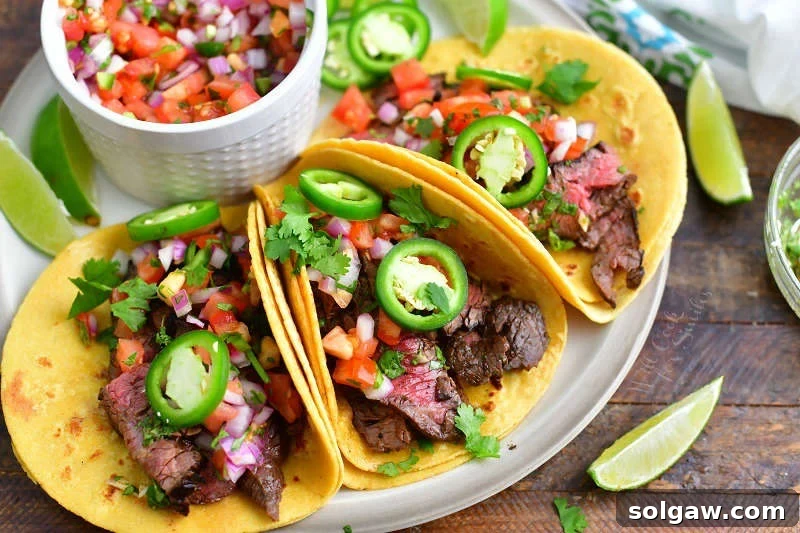 three steak tacos topped with jalapeno rings and pico on a light brown plate with a bowl of pico next to them and some lime slices around