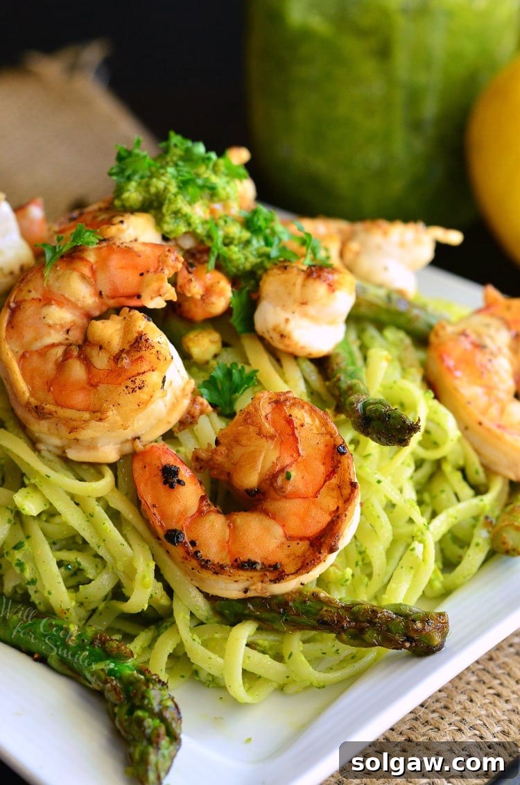 Vibrant lemon pistachio pesto pasta topped with succulent sauteed shrimp and fresh asparagus on a pristine white plate.