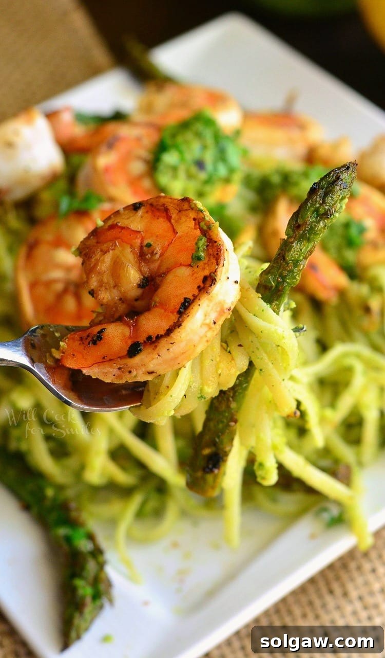 Close up of a fork holding sauteed shrimp and asparagus wrapped in pesto pasta over a white plate.