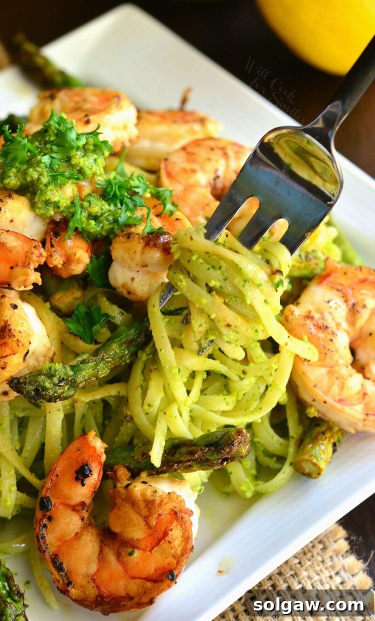 A fork swirling a generous portion of bright pesto pasta topped with shrimp and asparagus from a white plate.