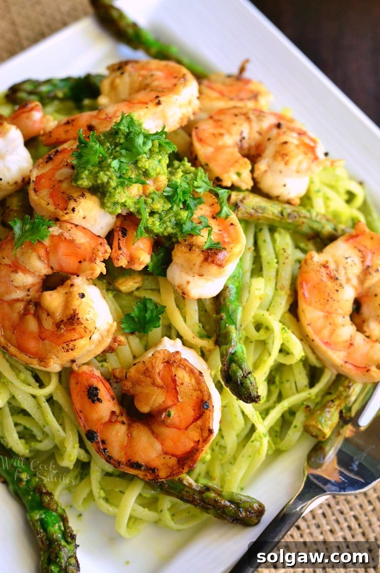 Bright pesto pasta topped with sauteed shrimp and asparagus on a white plate, ready to be served.