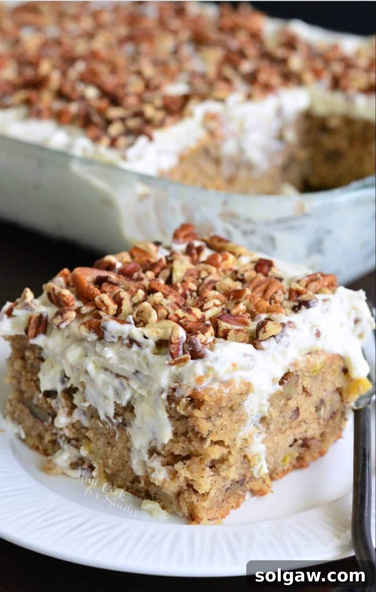 A square piece of delightfully moist Hummingbird Poke Cake on a white plate, showcasing the creamy frosting and a hint of the cake's texture. The baking dish is visible in the background.