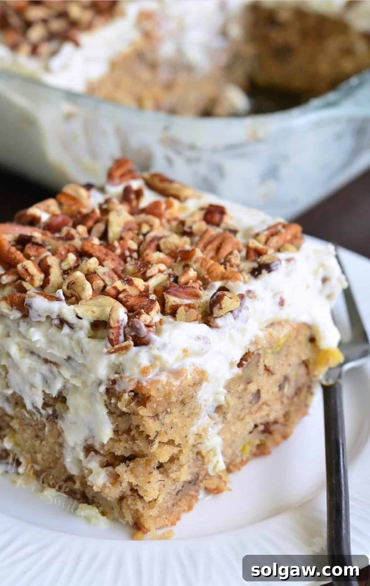 A delicious slice of Hummingbird Poke Cake on a white plate, garnished with nuts and accompanied by a fork, ready to be enjoyed.