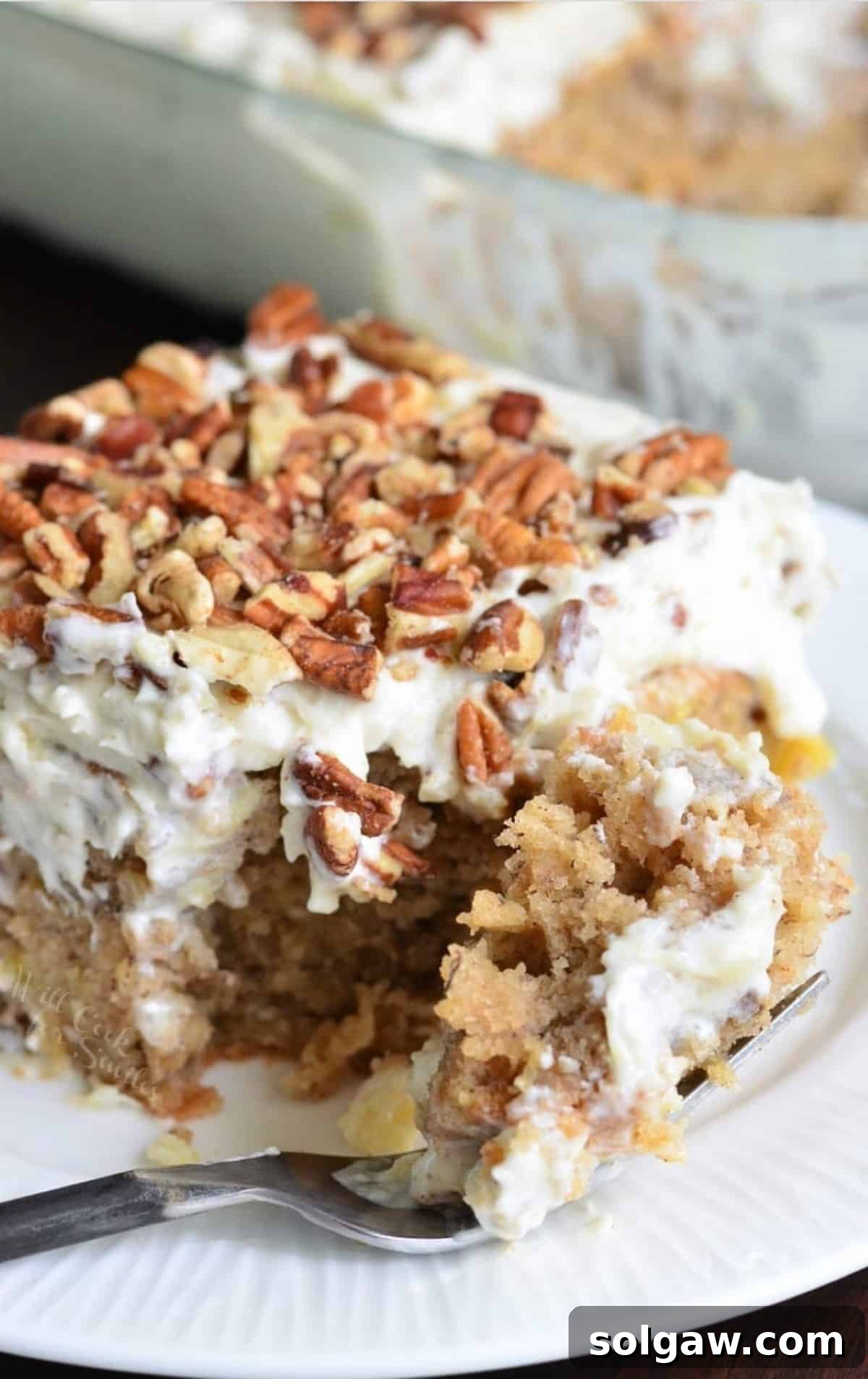 A delectable square slice of Hummingbird Poke Cake on a white plate, with a fork gently taking a piece, highlighting its moist texture and rich layers.