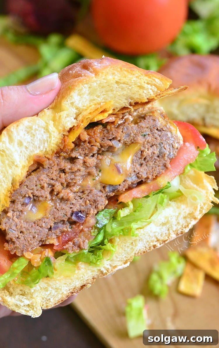 A hand holding a Stuffed Taco Burger, cut in half to showcase the melted cheese, with fresh lettuce and tomato on a bun, suggesting its deliciousness.