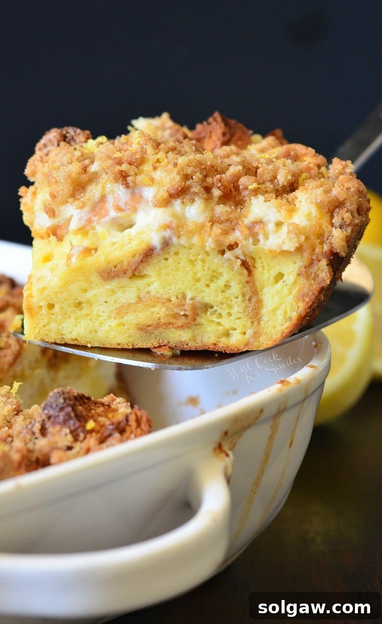 A close-up view of a perfectly baked slice of Lemon Cheesecake French Toast Casserole on a spatula, highlighting the creamy cheesecake layer nestled between brioche and topped with lemon crumble.
