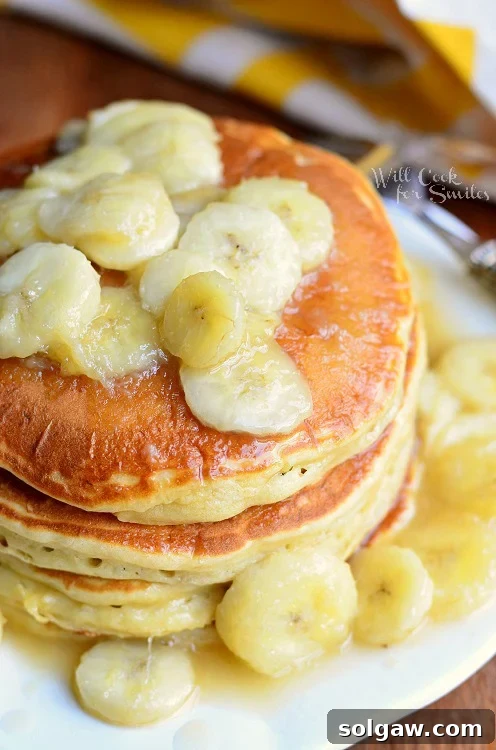 Overhead view of a beautiful stack of golden-brown Bananas Foster Pancakes on a pristine white plate, gracefully resting on a rustic wooden table with a soft yellow and white striped cloth artfully placed in the background.