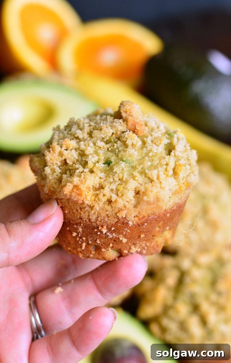 Close-up of a hand holding a freshly baked Avocado Banana Muffin with Orange Streusel, showcasing its beautiful crumb and topping texture.