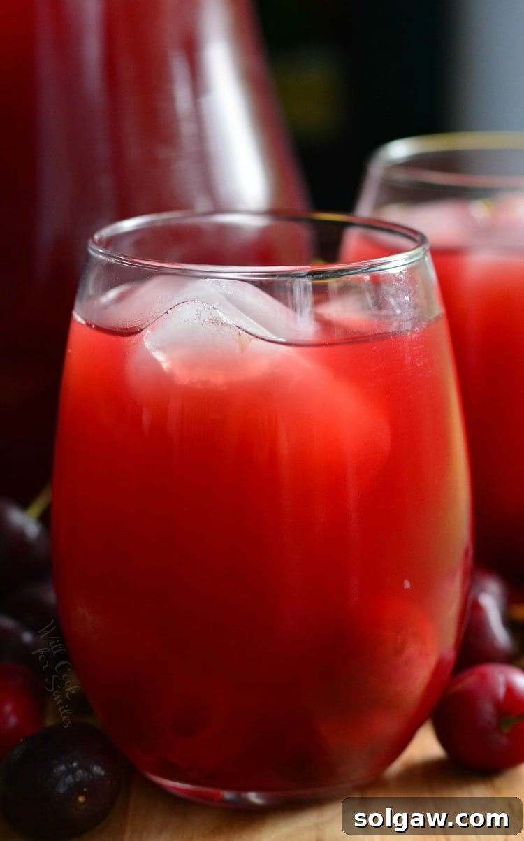 Homemade Cherry Arnold Palmer in a stemless wine glass on a table with a pitcher in the background and cherries around it 