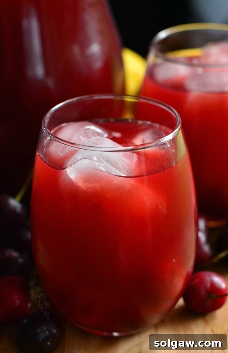 a pitcher and 2 glasses of Cherry Arnold Palmer on a cutting board with cherries around it 