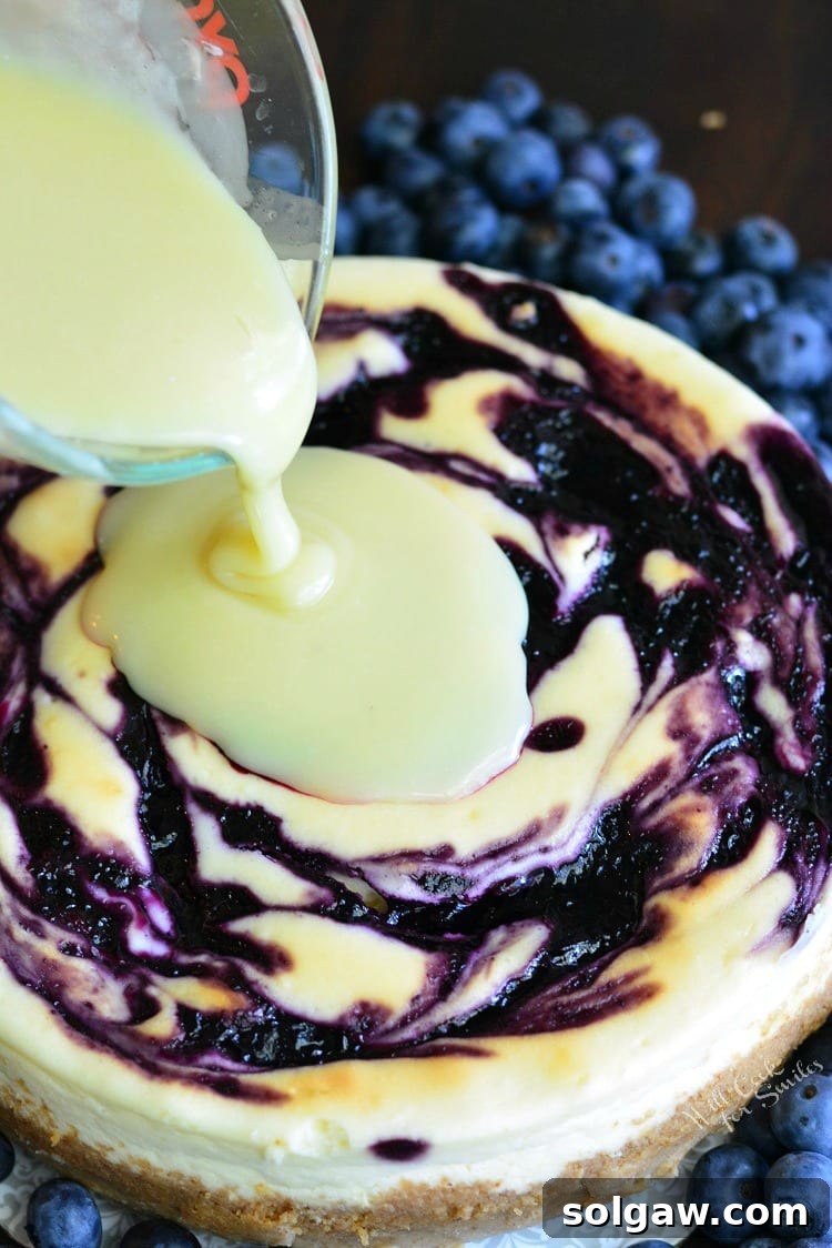White chocolate ganache being poured generously over a blueberry cheesecake, creating a smooth, glossy finish.