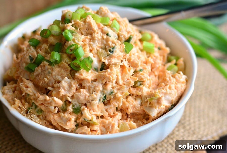 Zesty Buffalo Chicken Salad in a white bowl with green onions on top and a fork, placed on a rustic wooden table setting, inviting a delicious meal