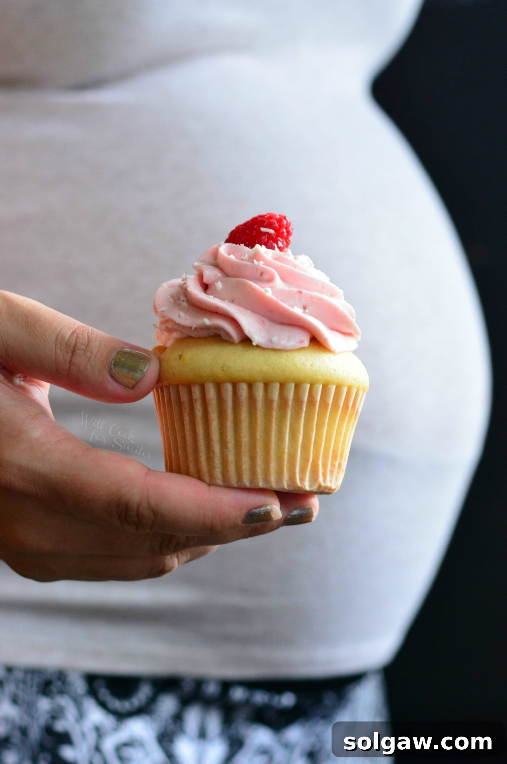 A hand gently holding a frosted cupcake with a fresh raspberry on top, positioned in front of a pregnant belly, signaling a special announcement.