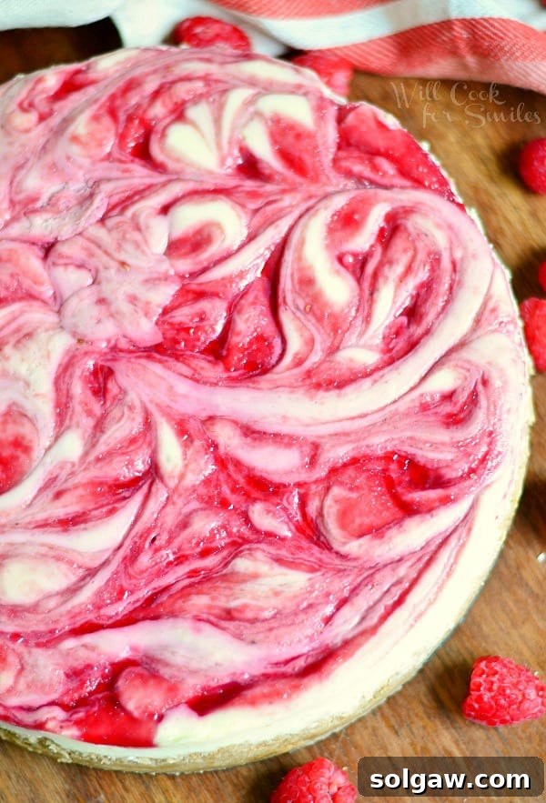 A whole white chocolate raspberry swirl cheesecake presented on a rustic wooden table, with fresh raspberries in the foreground and a soft white cloth in the background.