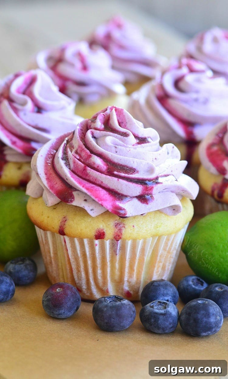 A vibrant key lime cupcake crowned with fluffy blueberry frosting, surrounded by fresh blueberries for a burst of color and flavor.