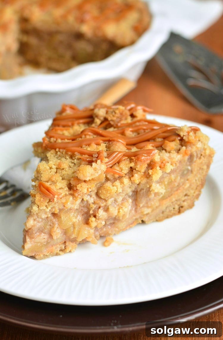 A delectable slice of Caramel Apple Cookie Pie resting on a pristine white plate, with the remainder of the golden-brown pie nestled in its pan, softly blurred in the background. The slice showcases distinct layers of cookie crust, apple filling, and streusel topping.
