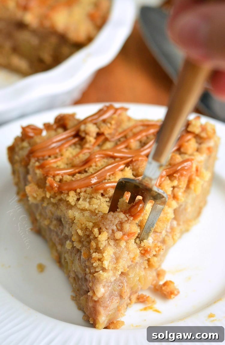 An overhead shot of the Caramel Apple Cookie Crust Pie, baked to golden perfection, resting on a white plate with a fork poised nearby, ready for the first bite. The texture of the streusel topping and the richness of the caramel are visible.