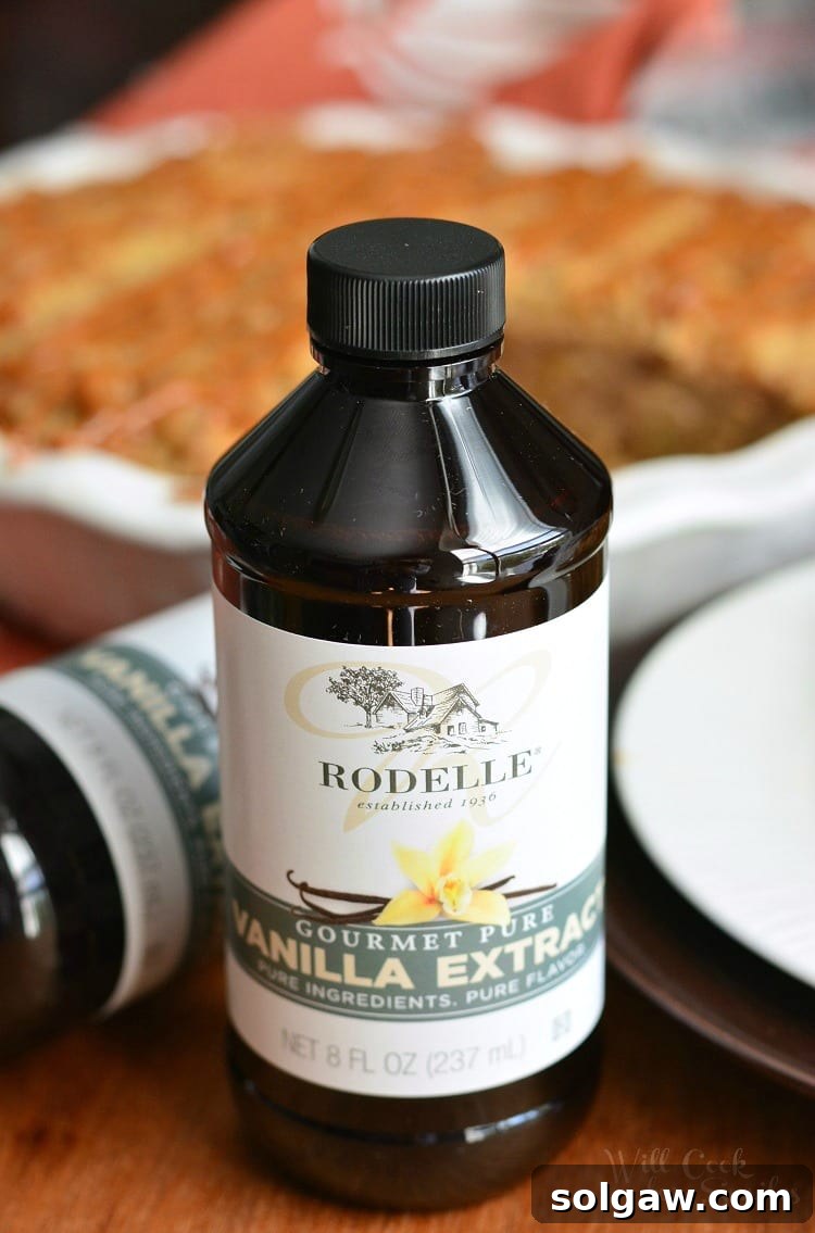A bottle of high-quality Rodelle vanilla extract stands prominently in the foreground, with the inviting Caramel Apple Cookie Crust Pie softly blurred in the background, hinting at the key ingredient that elevates the pie's flavor.