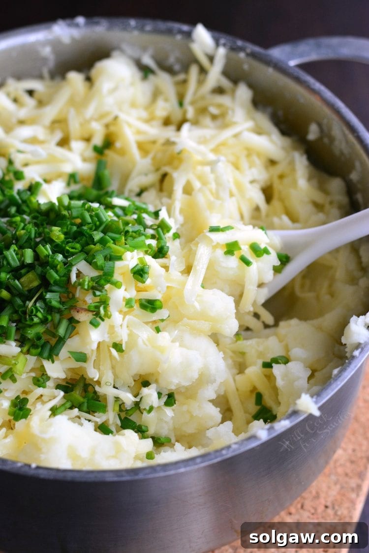 Large pot with creamy White Cheddar and Chive Mashed Potatoes, garnished with grated cheese and fresh chives, and a plastic spoon