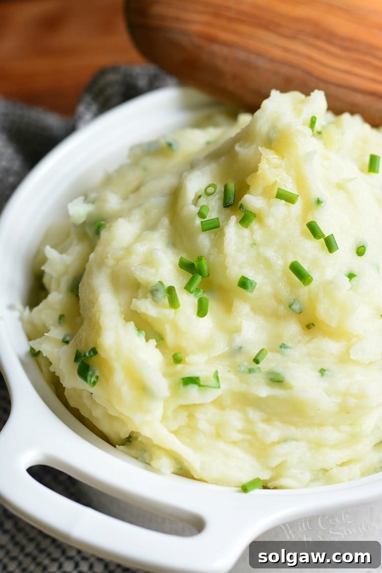 White serving bowl filled with a generous portion of White Cheddar and Chive Mashed Potatoes, with a wooden spoon scooping out a portion