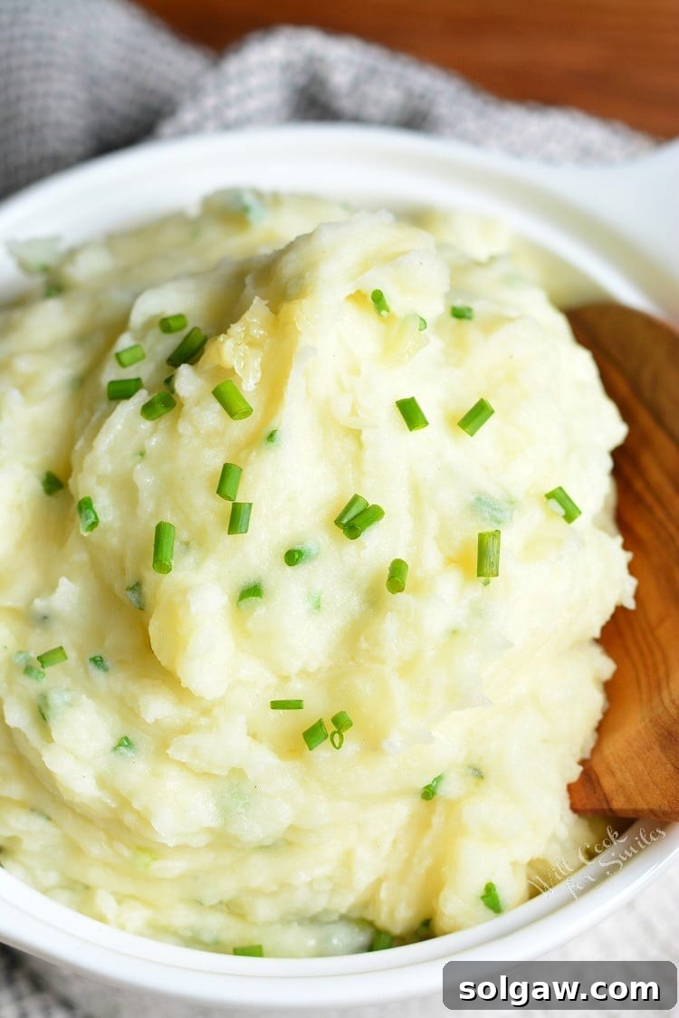 Serving bowl of creamy White Cheddar and Chive Mashed Potatoes, garnished with fresh chives, next to a wooden spoon