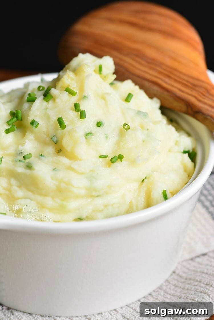 White bowl of fluffy White Cheddar and Chive Mashed Potatoes, topped with chopped fresh chives, and a wooden spoon