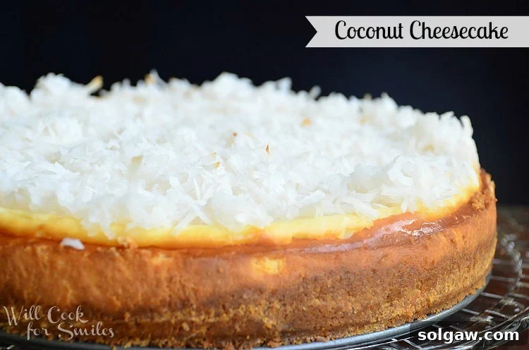 The full, round coconut cheesecake, with a single slice removed, elegantly presented on a glass cake platter. The rich coconut topping is clearly visible, inviting a taste of its tropical sweetness.